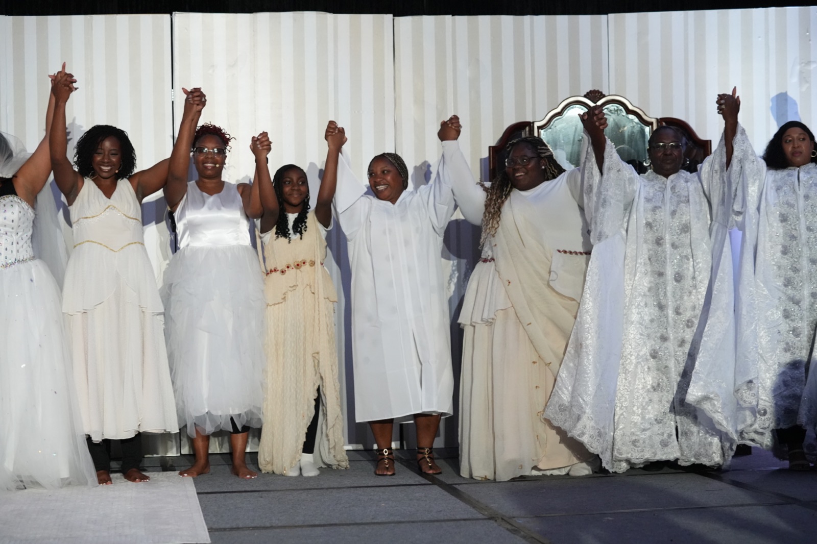 Company members in white and gold raise their joined hands at the curtain call