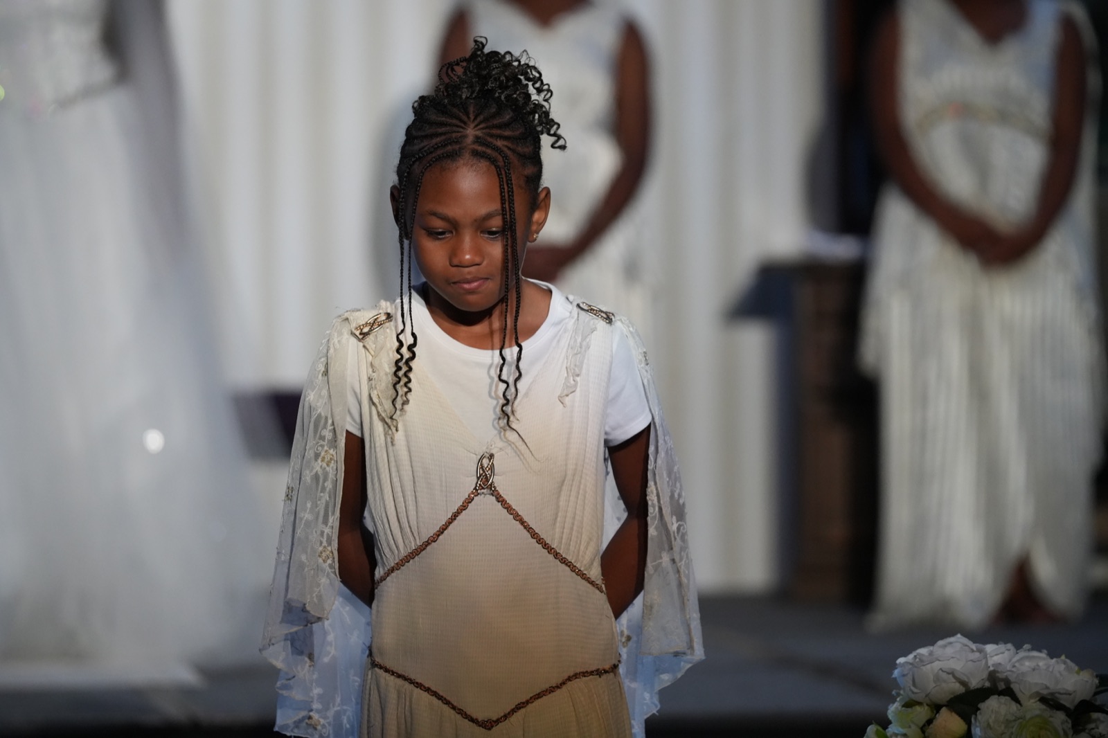 A young performer in cream and gold stands before the white-clad company